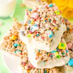 Lucky Charms Treats arranged on a white plate with a glass of milk and daffodils in the background.