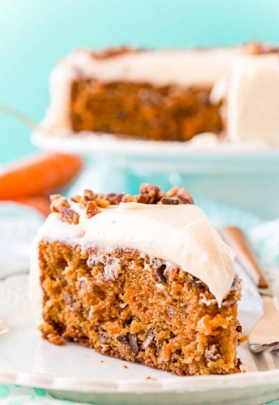 Slice of carrot cake on a white plate with a fork. Full cake on a cake stand in the background.