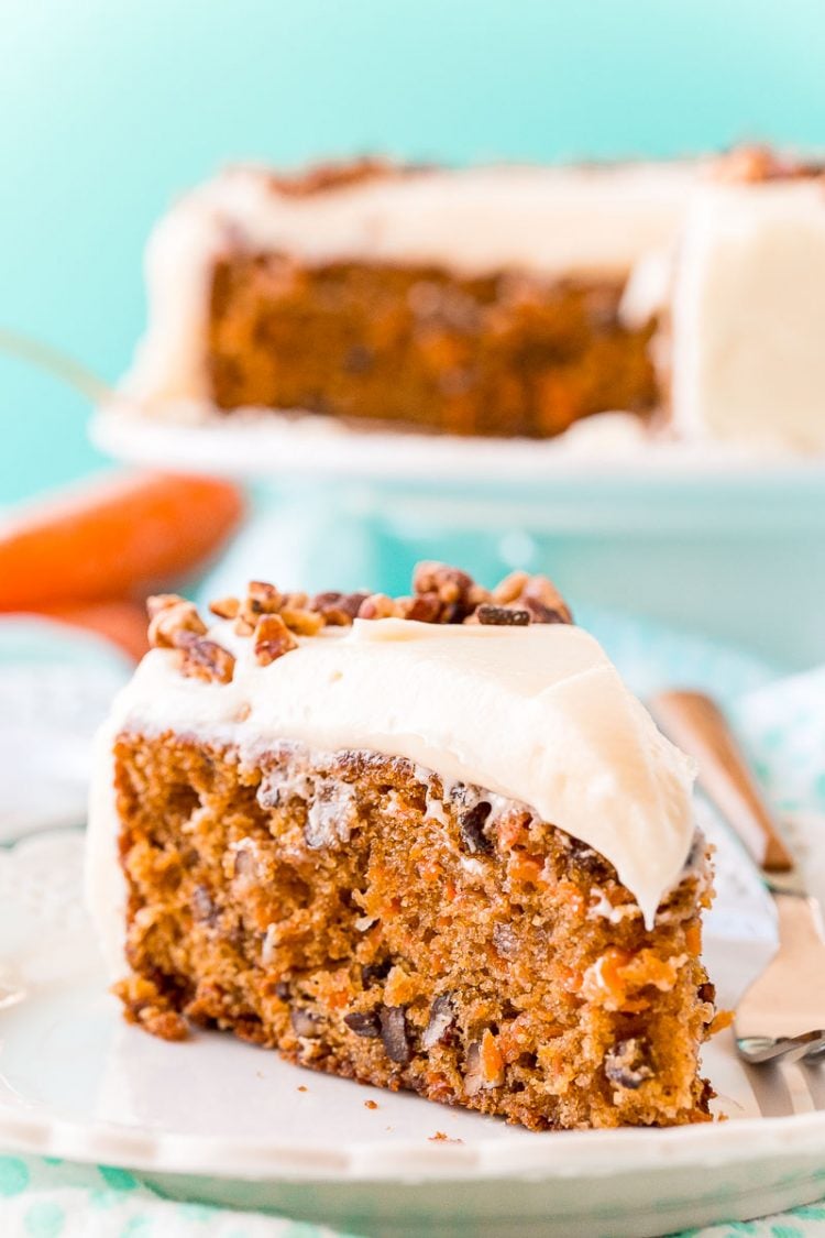 Slice of carrot cake on a white plate with a fork. Full cake on a cake stand in the background.