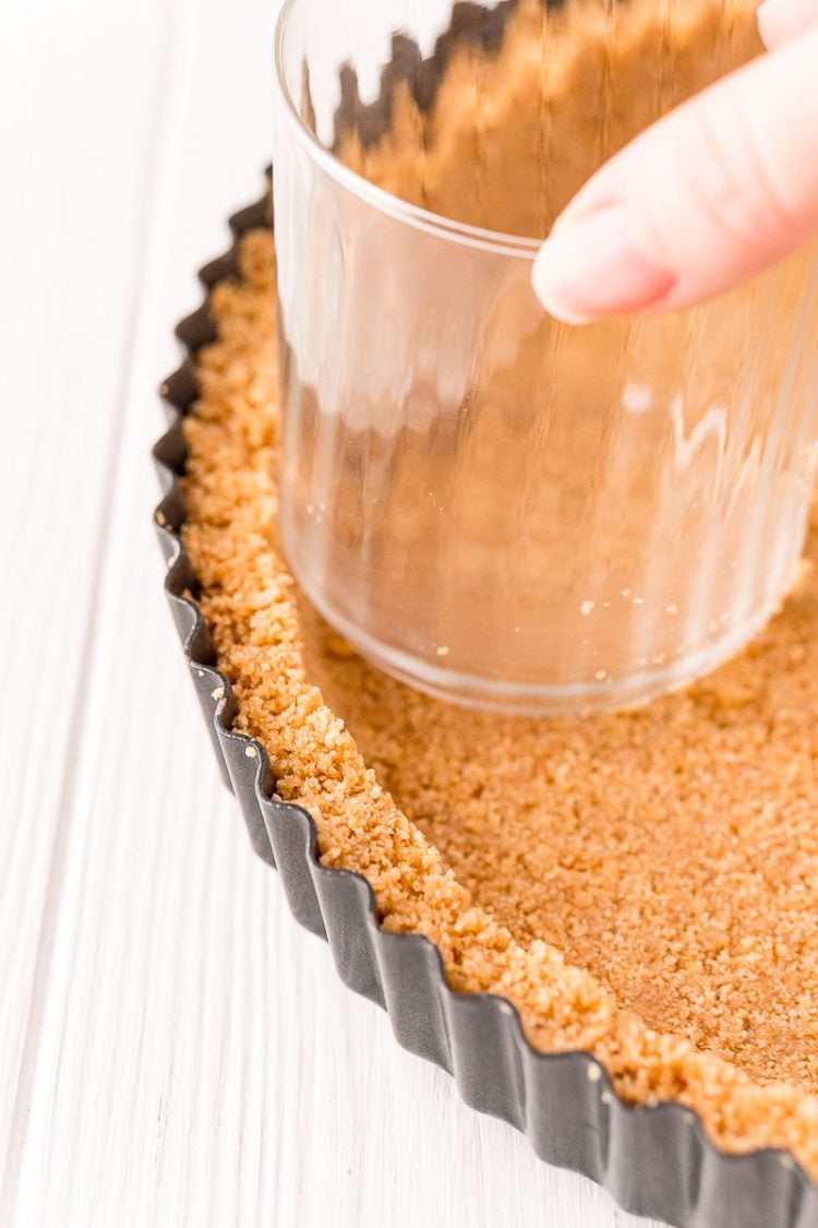Woman's hand using a glass to shape a graham cracker crust in a tart pan.