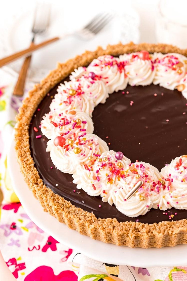 Chocolate tart topped with whipped cream and sprinkles on a white cake stand.
