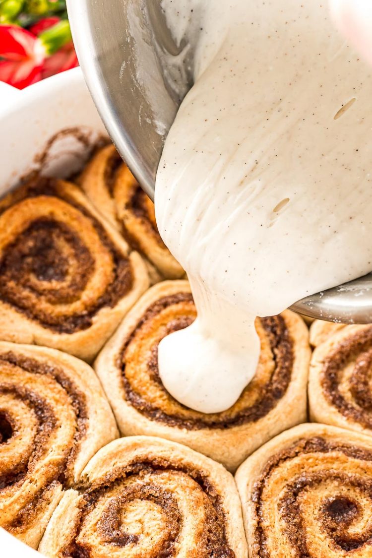 Baked cinnamon rolls in a casserole dish being iced with vanilla icing.