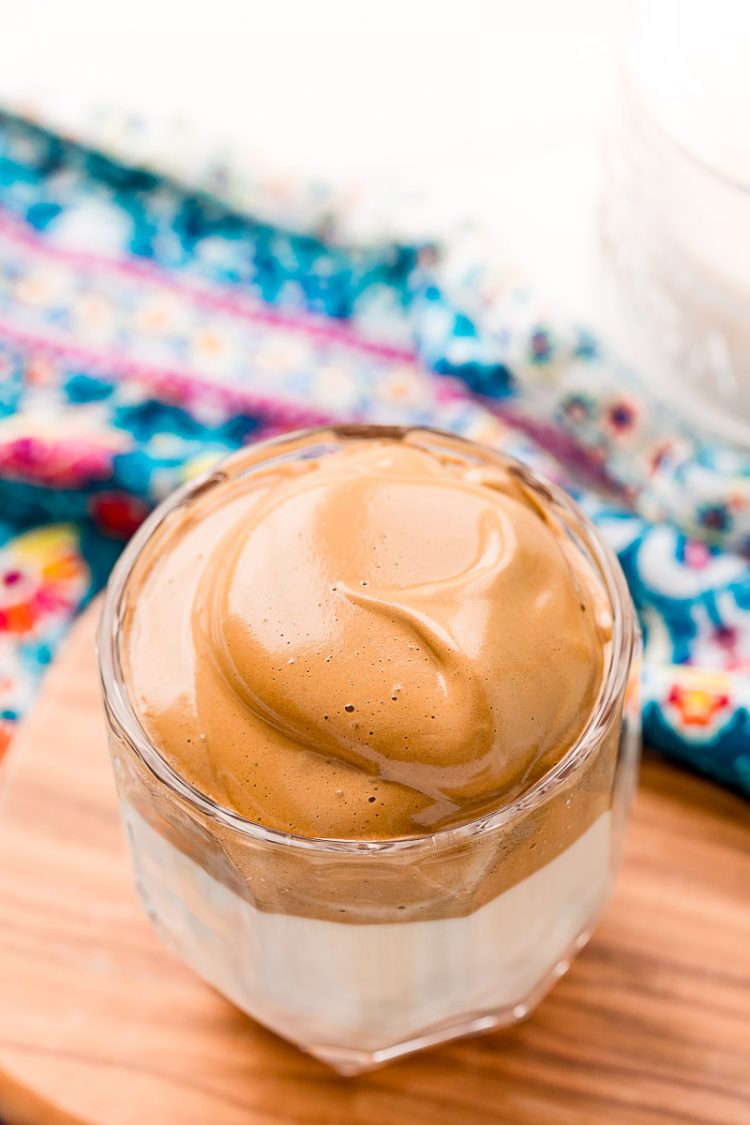 Angled overhead photo of a glass of whipped Dalgona coffee on a small cutting board.