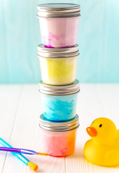 Small jars of paint stacked on top of each other with a rubber ducky and paint brushes sitting next to them on a white table.