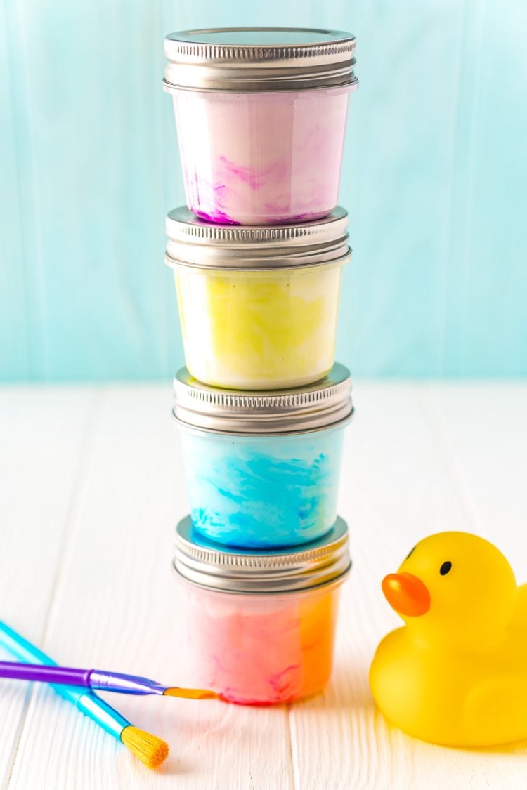 Small jars of paint stacked on top of each other with a rubber ducky and paint brushes sitting next to them on a white table.