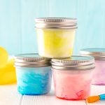 Small jars of paint stacked on top of each other with a rubber ducky and paint brushes sitting next to them on a white table.
