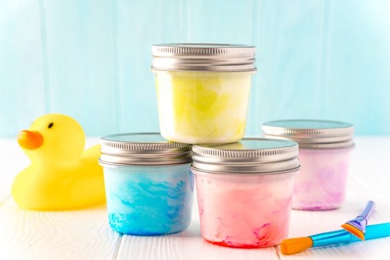 Small jars of paint stacked on top of each other with a rubber ducky and paint brushes sitting next to them on a white table.