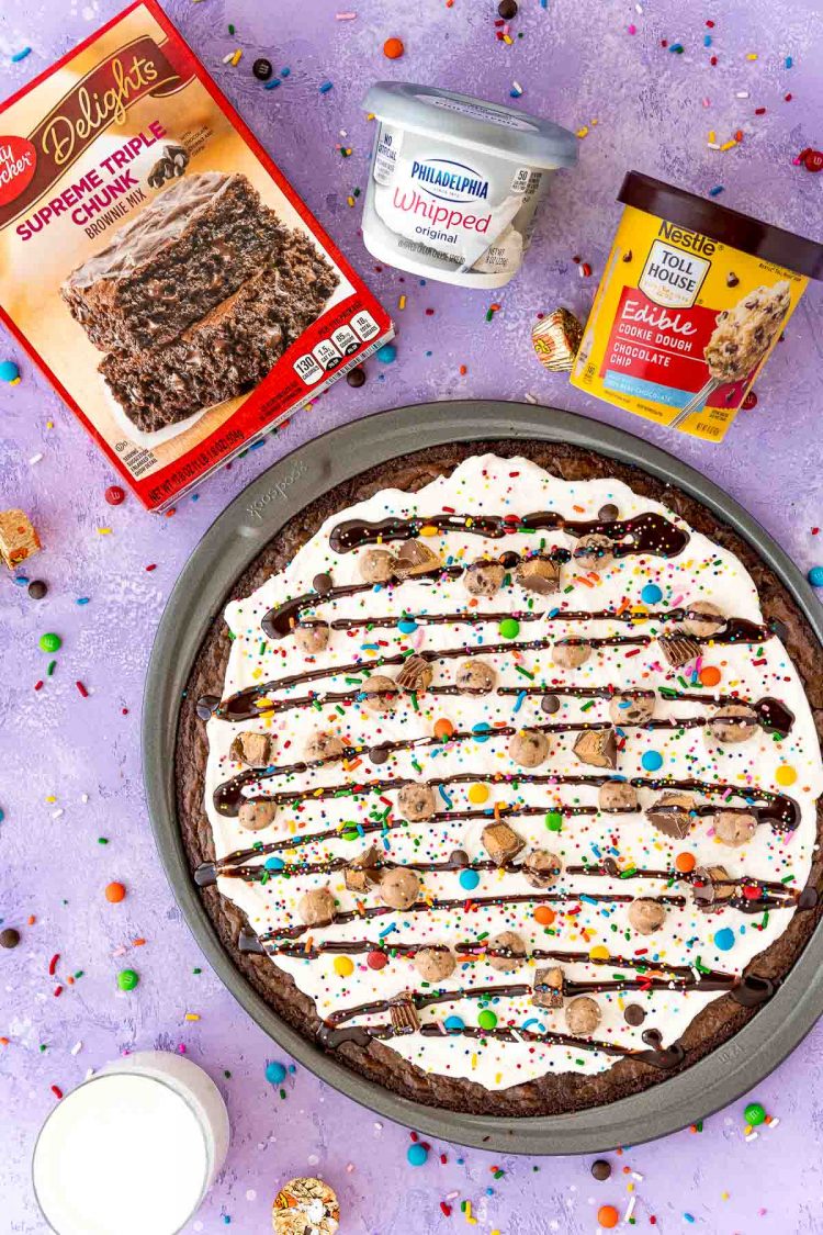 Overhead photo of brownie pizza with a box of brownie mix, container of cream cheese, and container of edible cookie dough.