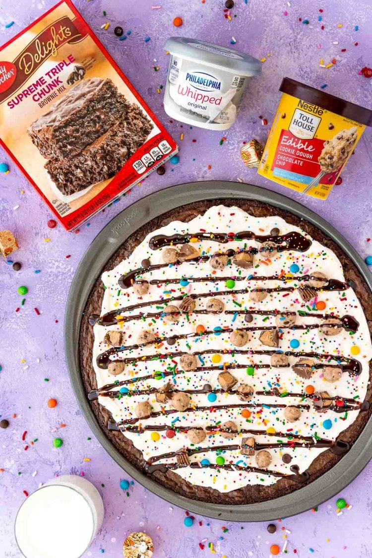 Overhead photo of brownie pizza with a box of brownie mix, container of cream cheese, and container of edible cookie dough.