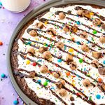 Close up photo of brownie dessert pizza in a pan on a purple surface with candy scattered around.