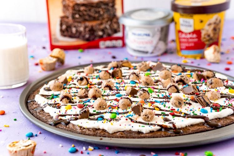 Brownie pizza on a pan with ingredients and a glass of milk in the background.