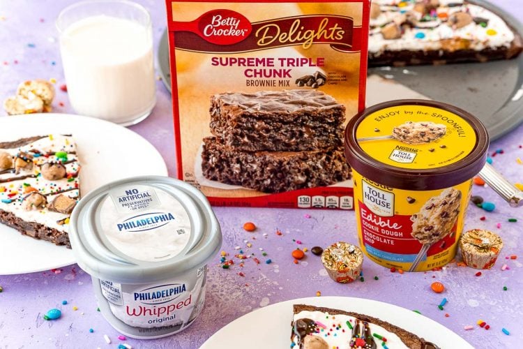 Ingredients to make brownie pizza on a purple surface surrounded by candy and brownie pizza slices on white plates.