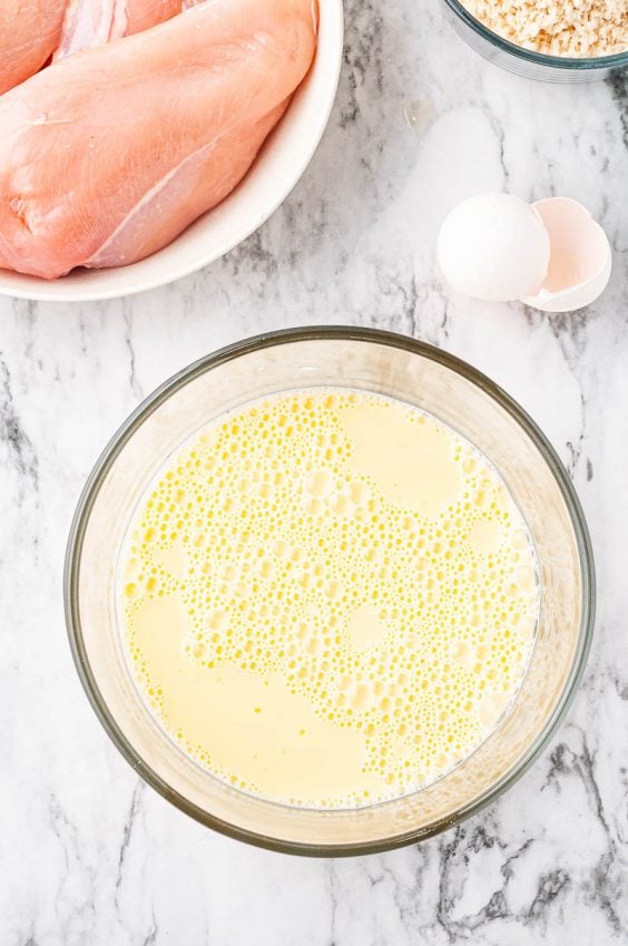 Milk and eggs beat together in a glass bowl with raw chicken sitting next to it on a marble countertop.