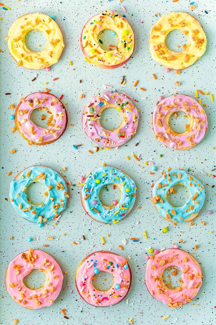 Overhead photo of apple slices with colorful frosting on them on a blue baking sheet with sprinkles.