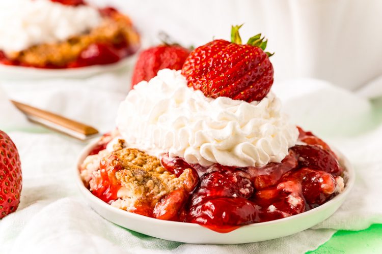 Close up photo of strawberry cobbler on a white plate topped with whipped cream and fresh strawberry.