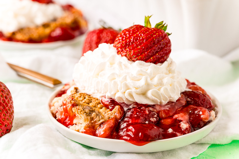 Close up photo of strawberry cobbler on a white plate topped with whipped cream and fresh strawberry.