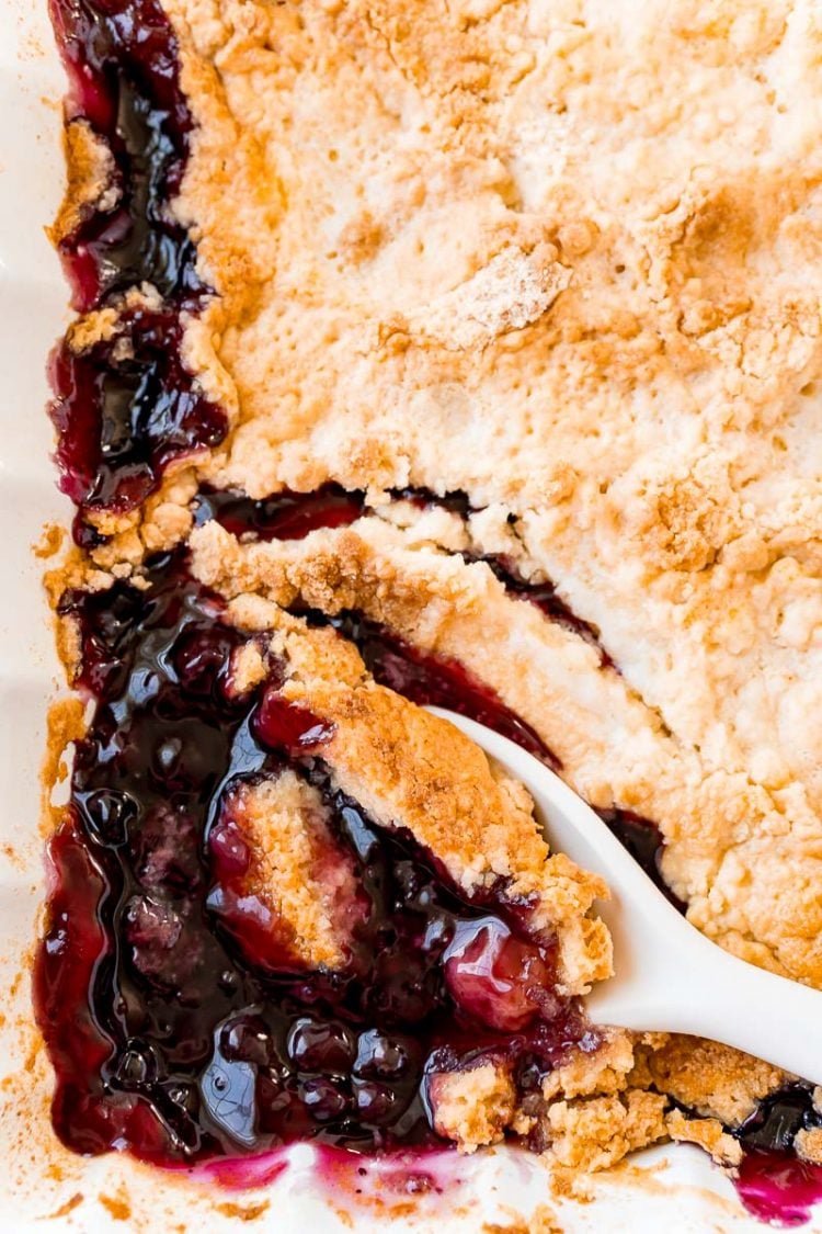 Overhead photo of a white serving spoon scooping bluberry dump cake cubbler out of a white baking dish.