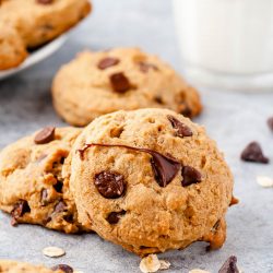 Chocolate Chip cookies on a counter with chocolate chips scattered around and a glass of milk in the background.