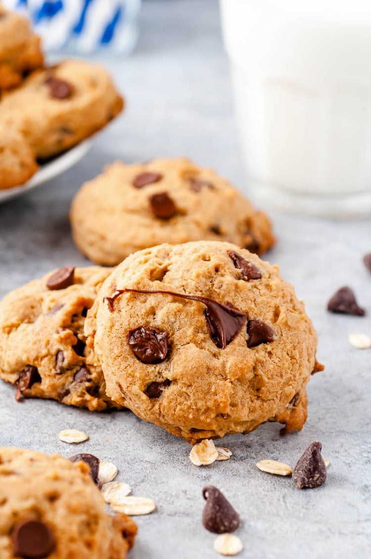 Chocolate Chip cookies on a counter with chocolate chips scattered around and a glass of milk in the background.