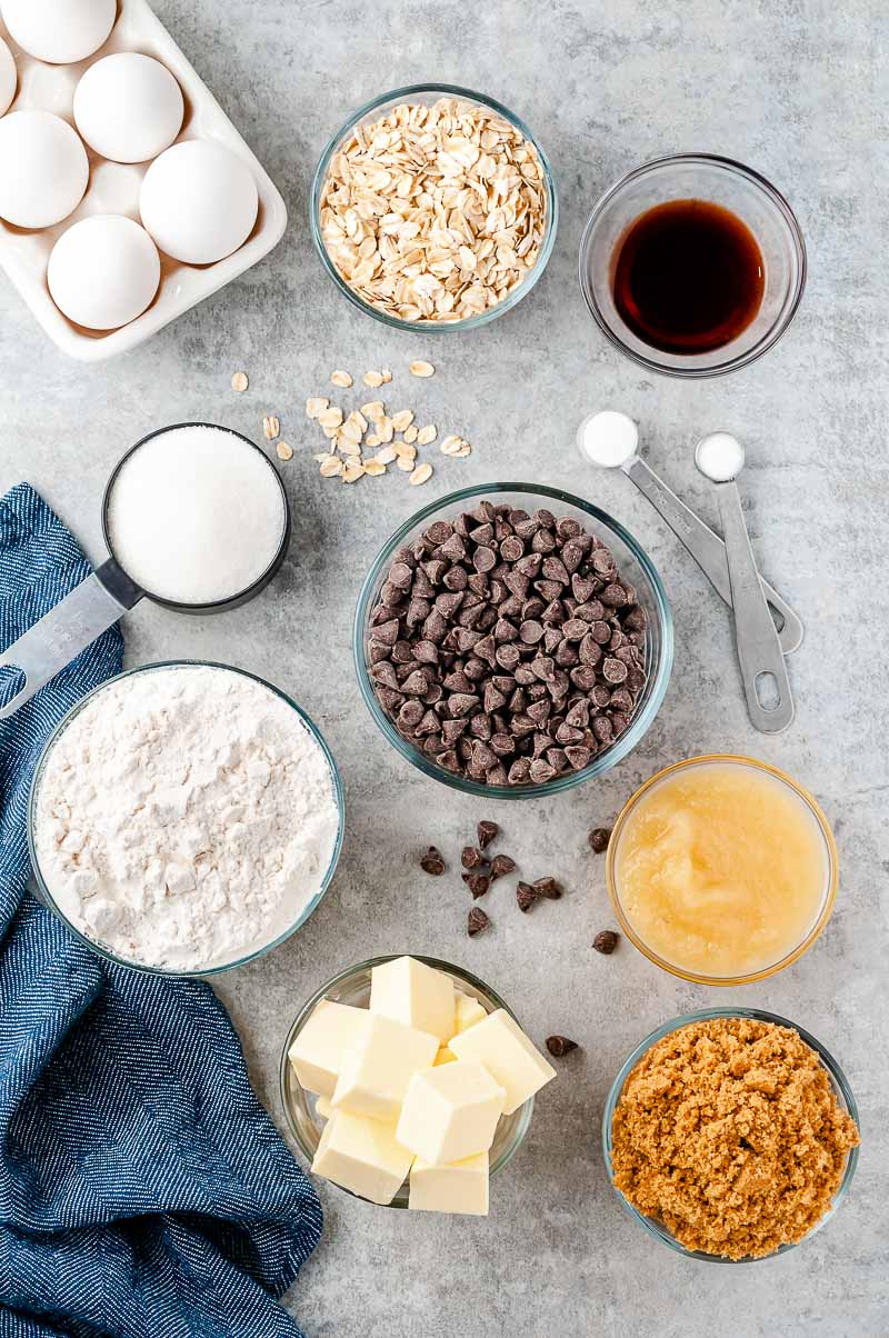 Ingredients to make Oatmeal Chocolate Chip Cookies in prep bowls on a grey counter with a blue napkin.