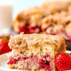 A slice of strawberry coffee cake on a small white plate with a strawberry sitting next to it. More coffee cake in the background.