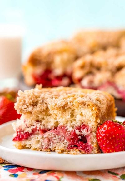 A slice of strawberry coffee cake on a small white plate with a strawberry sitting next to it. More coffee cake in the background.