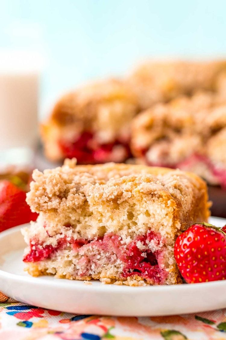 A slice of strawberry coffee cake on a small white plate with a strawberry sitting next to it. More coffee cake in the background.