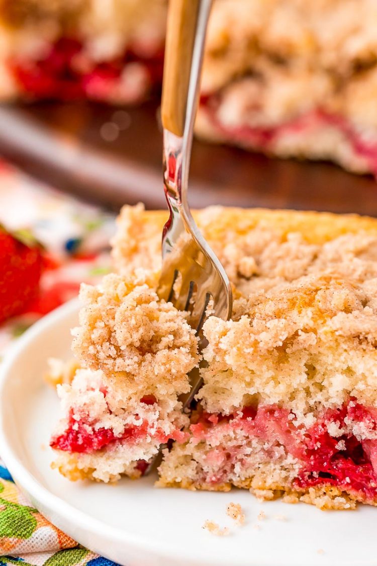 A fork taking a bite out of a strawberry coffee cake slice on a white plate.