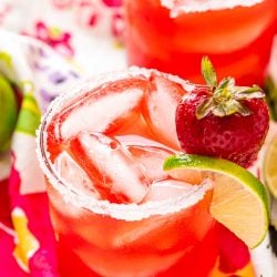 Glass filled with a strawberry cocktail on a colorful napkin with strawberries and limes scattered around it.