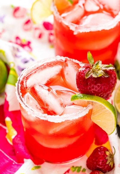 Glass filled with a strawberry cocktail on a colorful napkin with strawberries and limes scattered around it.