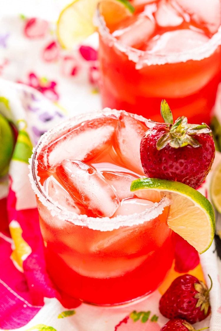 Glass filled with a strawberry cocktail on a colorful napkin with strawberries and limes scattered around it.