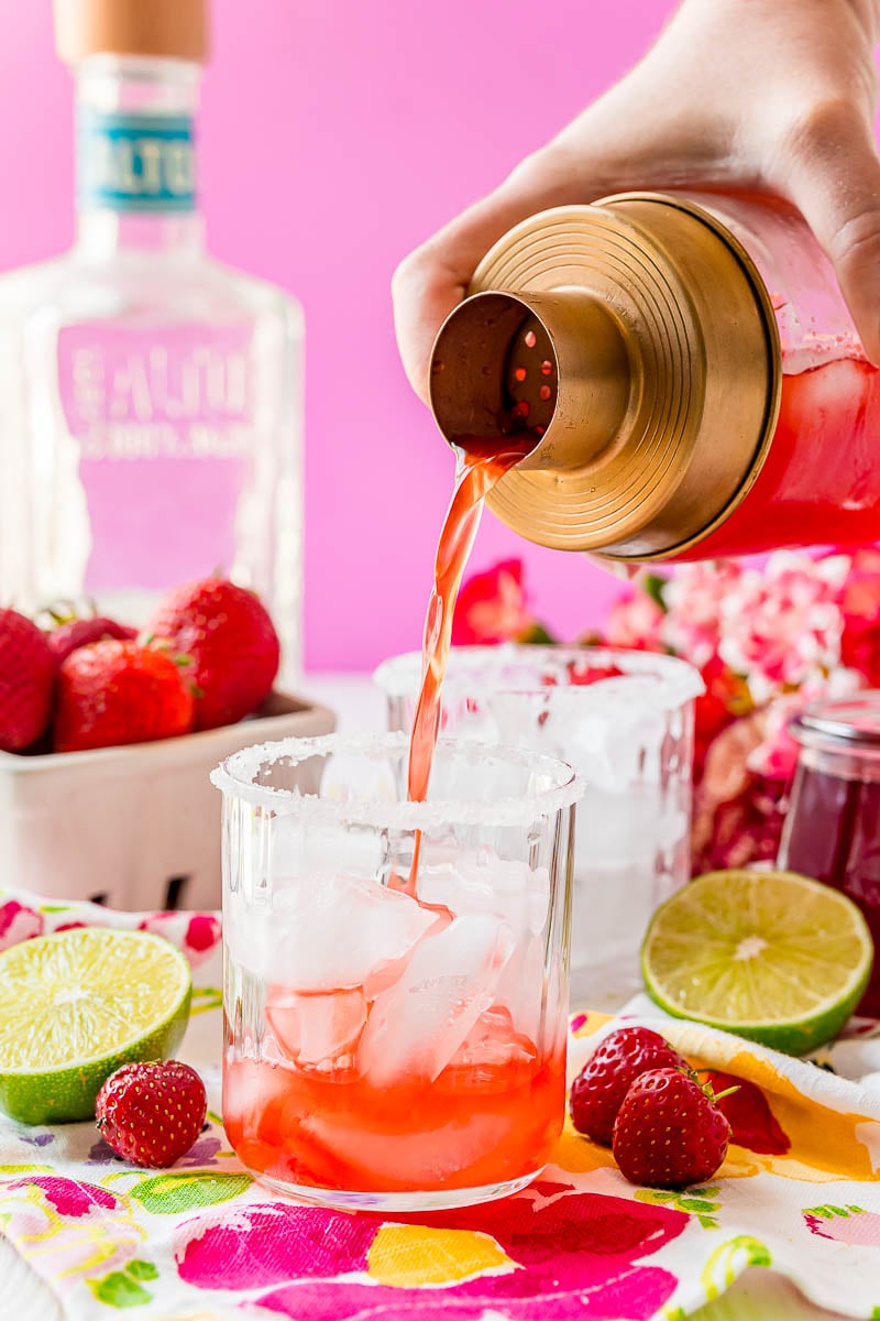Strawberry margarita being poured out of a cocktail shaker into a glass with ice.