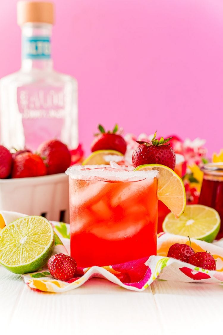 Straight on photo of a strawberry margarita on the rocks garnished with lime wedge and strawberry. Bottle of tequila in the background.