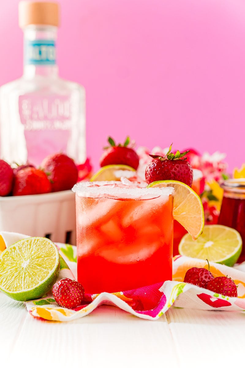 Straight on photo of a strawberry margarita on the rocks garnished with lime wedge and strawberry. Bottle of tequila in the background.