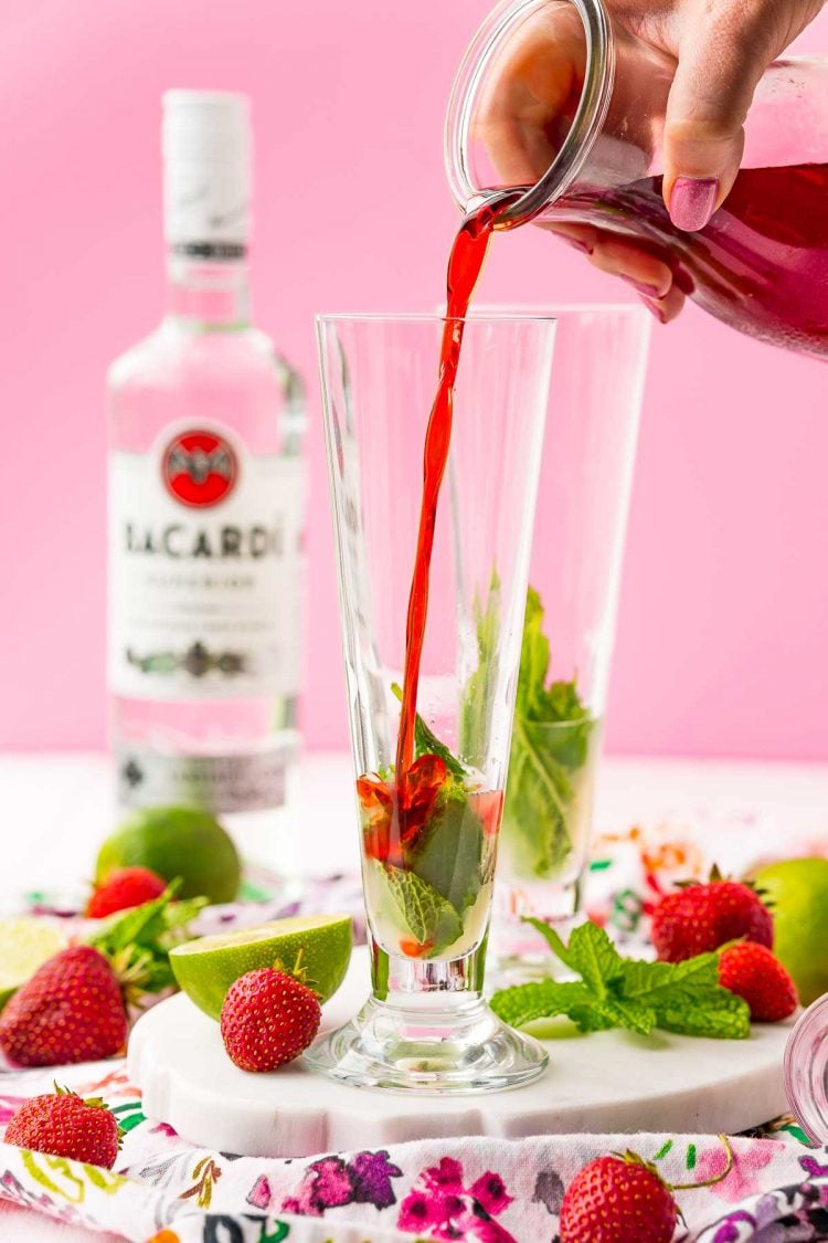 Strawberry simple syrup being poured into a tall cocktail glass with lime juice and mint.