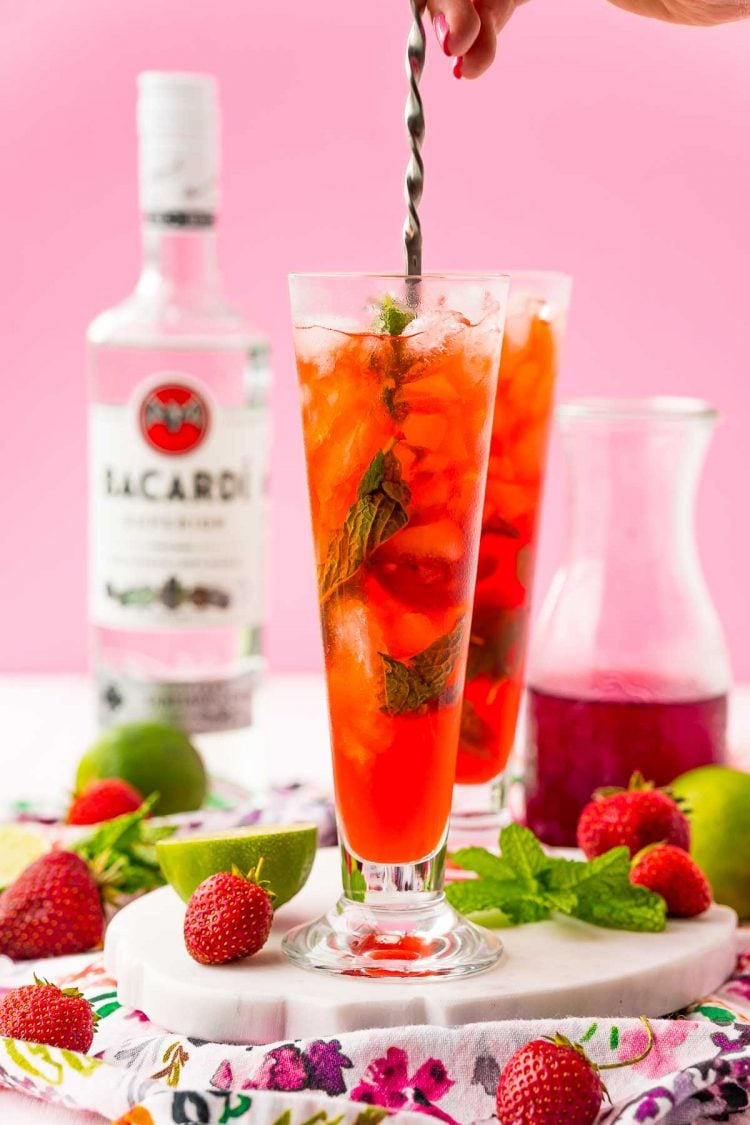 A woman's hand holding a bar spoon to mix a red cocktail in a tall glass.