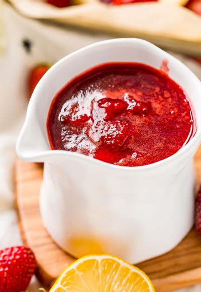 Strawberry sauce in a white serving cup with strawberries and lemons scattered around it.