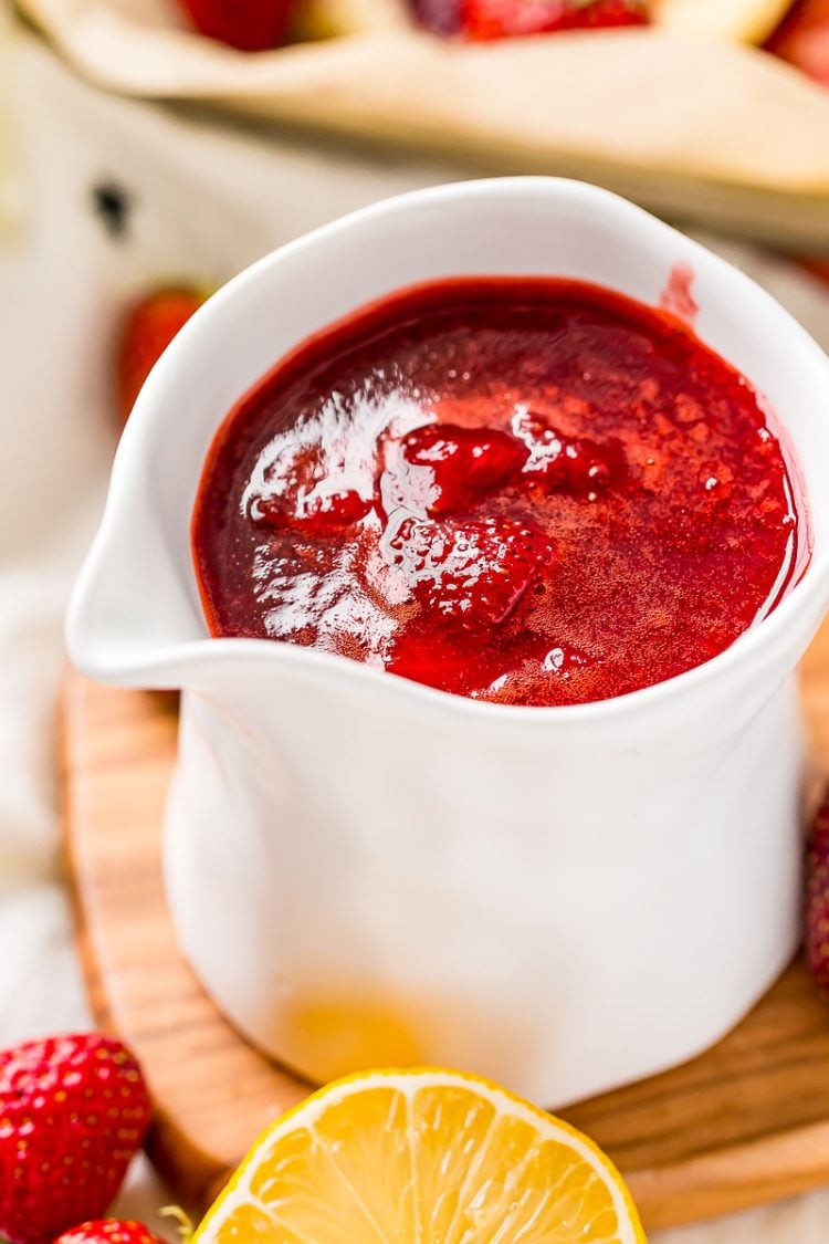 Strawberry sauce in a white serving cup with strawberries and lemons scattered around it.