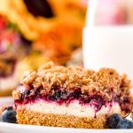 Close up photo of a slice of blueberry cheesecake bars on a white plate with milk and sunflowers in the background.