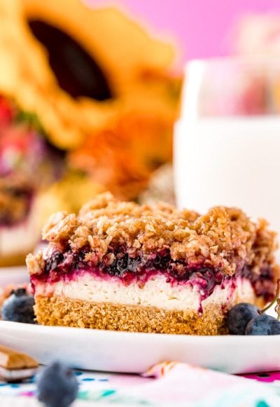 Close up photo of a slice of blueberry cheesecake bars on a white plate with milk and sunflowers in the background.