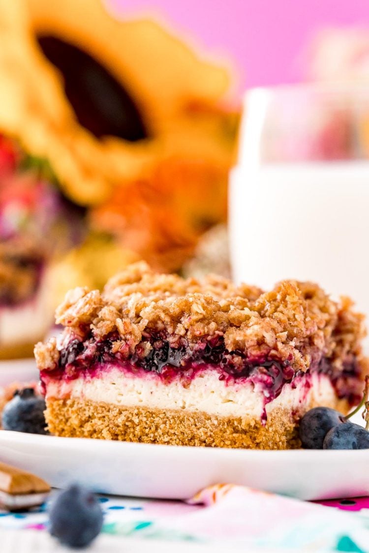 Close up photo of a slice of blueberry cheesecake bars on a white plate with milk and sunflowers in the background.