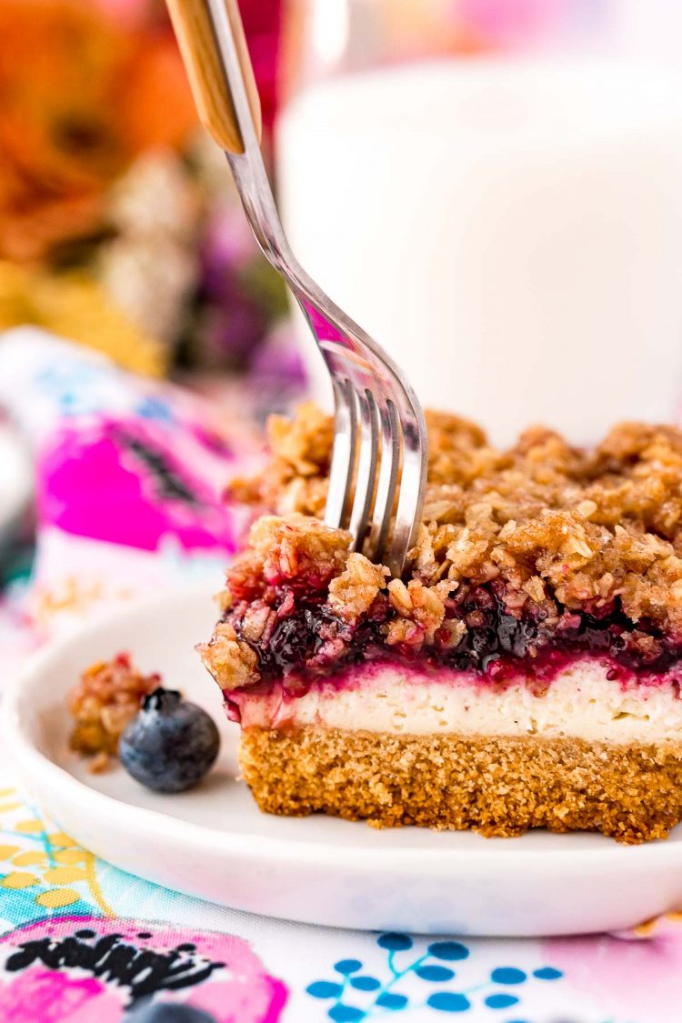 Fork taking a bite out of a slice of blueberry cheesecake with crumb topping.