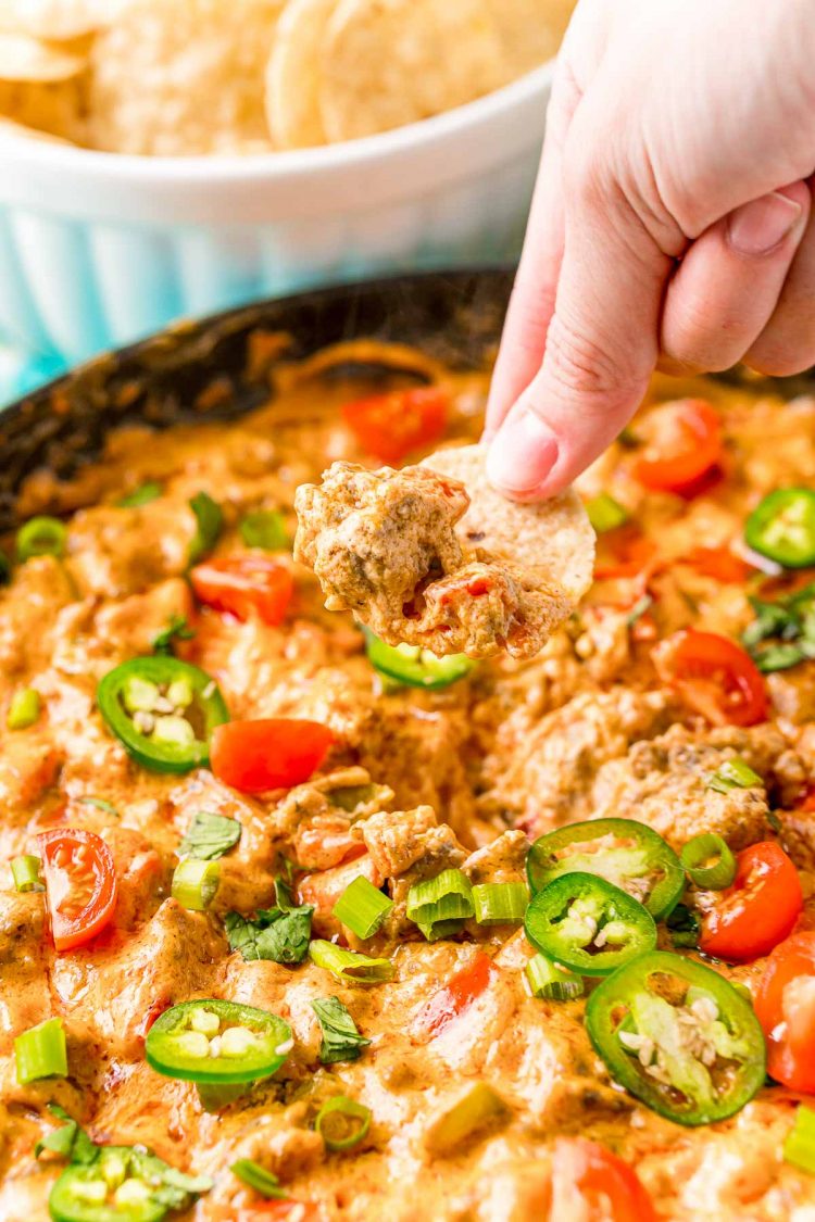 A woman's hand pulling a tortilla chip with cheesy dip out of a bowl of dip.