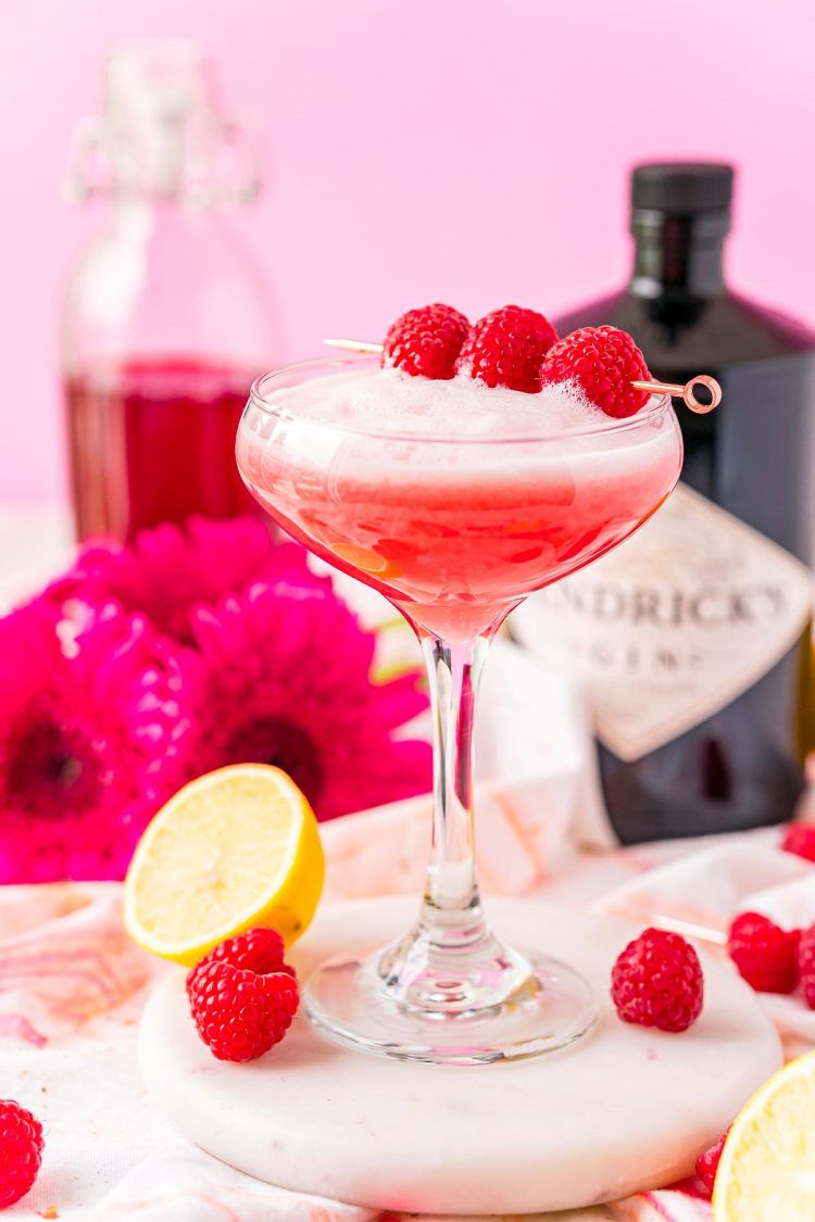 Clover club cocktail on a white marble surface with pink flowers and bottles of simple syrup and gin in the background.