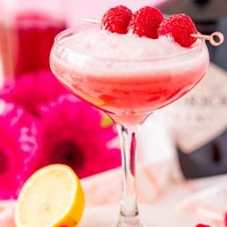 A cocktail glass filled with a pink foamy cocktail garnished with raspberries.