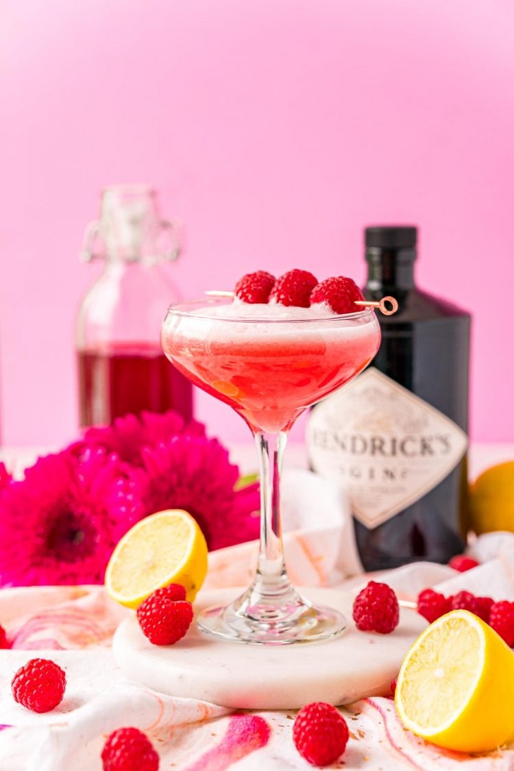 Clover club cocktail surrounded but lemon halves and raspberries with a bottle of gin in the background.