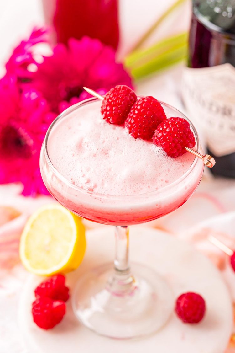 A cocktail glass filled with a pink foamy cocktail garnished with raspberries.