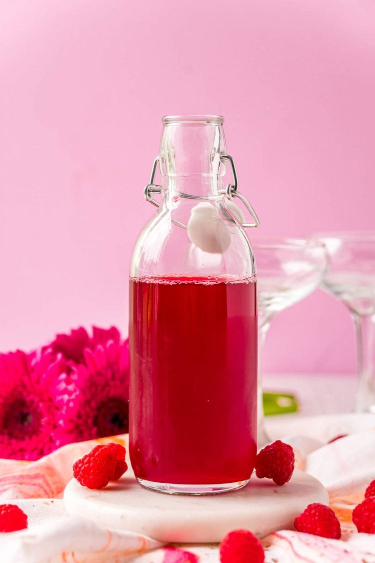 Raspberry simple syrup in a glass bottle surrounded by raspberries and pink flowers.