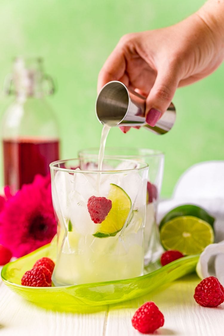 Lime juice being poured into a glass of ice with lime wedges and raspberries in it.