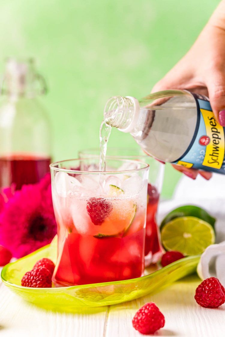 Club soda being added to a glass with lime juice and raspberry simple syrup in it.
